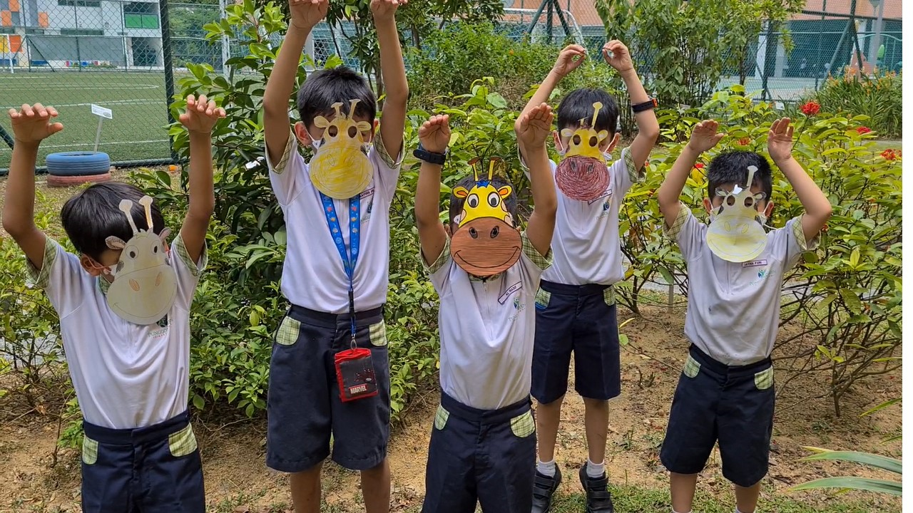 Five children in school uniforms wear handmade giraffe masks and raise their arms outdoors near green bushes.